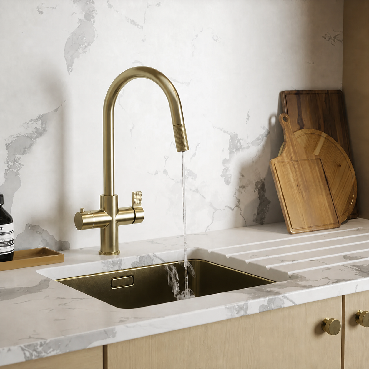A modern kitchen sink setup with a tall brushed brass gooseneck faucet running water into a matching brass undermount sink. The countertop is white marble with gray veining, and wooden cutting boards lean against the backsplash on the right.